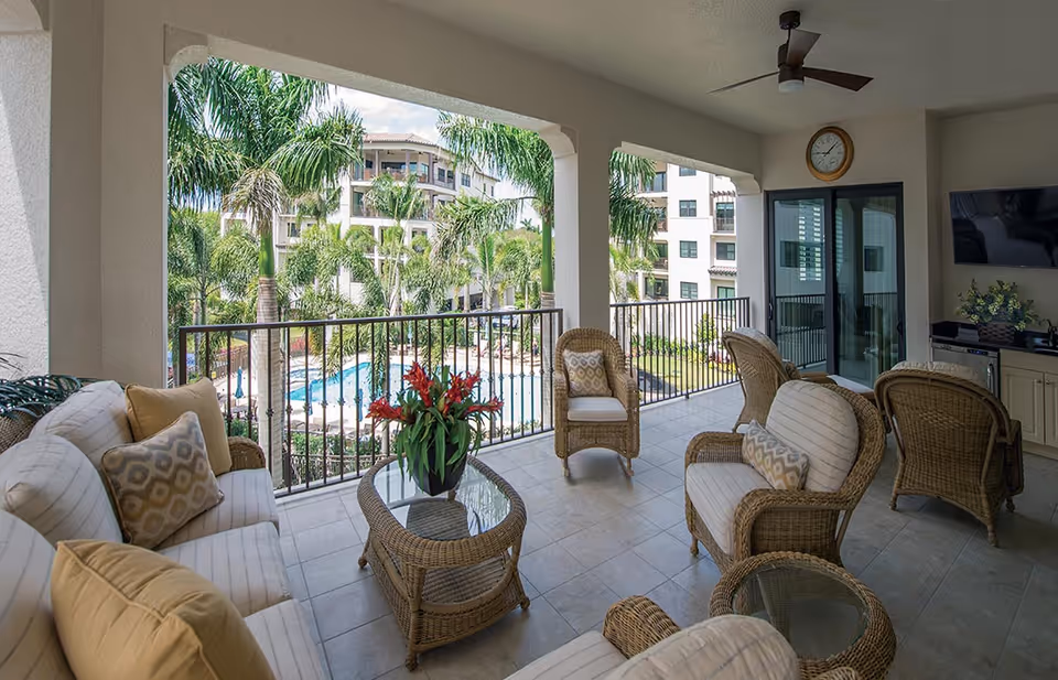 Covered balcony with wicker seating and a glass-top table overlooking palm trees and a swimming pool.