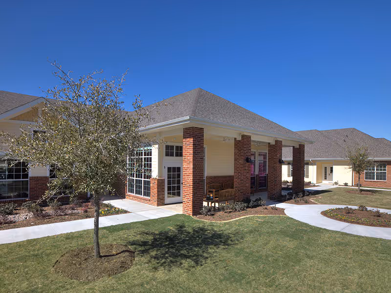 Exterior view of a senior living facility building with brick columns and beige siding under a clear blue sky. There is a small tree and landscaped grass area in the foreground with a concrete walkway leading to the entrance.