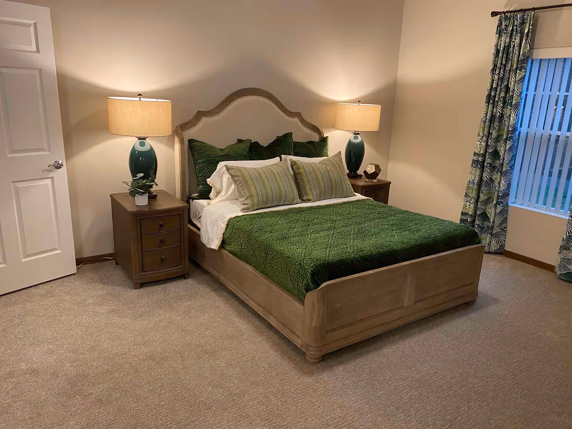 A neatly made bed with green bedding and multiple pillows in a bedroom. There are two wooden nightstands on either side of the bed, each with a green lamp. A window with patterned curtains is visible on the right side, and a closed white door is on the left. The room has beige walls and carpeted flooring.