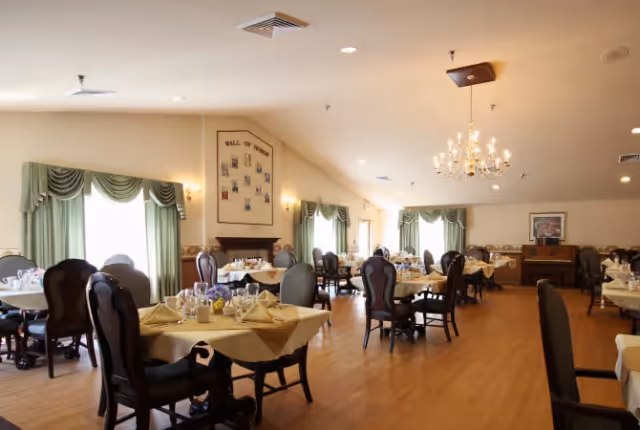 A spacious formal dining room with tables set with tablecloths and place settings, high-backed chairs, chandeliers, and green drapery.