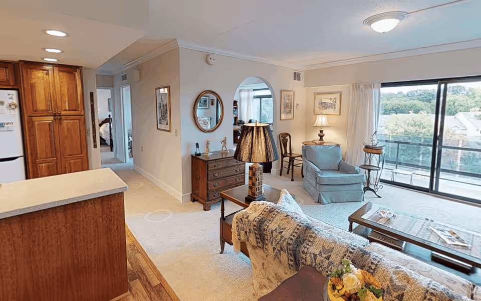 Interior view of a senior living facility showing a cozy living room with a blue armchair, a wooden coffee table, a lamp on a side table, and a large sliding glass door leading to a balcony with outdoor seating and a view of trees. Adjacent to the living room is a kitchen area with wooden cabinets and a refrigerator. A hallway leads to a bedroom visible in the background.