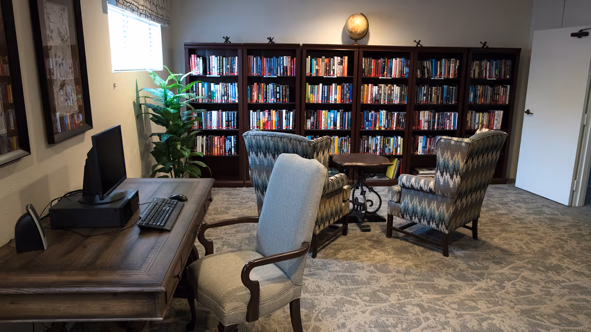 Small library-style common room with a desk and computer in the foreground, patterned armchairs around a side table, and bookshelves along the back wall.