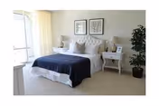 Bright bedroom with a neatly made bed topped by a dark blue throw, matching nightstands with lamps, framed art above and a potted plant by a window.