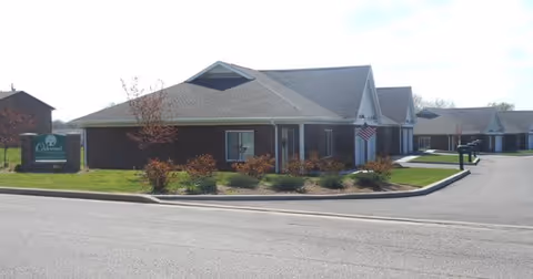 Exterior view of Oakwood Health Campus, a single-story brick building with a gray roof, surrounded by a well-maintained lawn and small shrubs. There is a sign with the facility name near the street and an American flag near the entrance.
