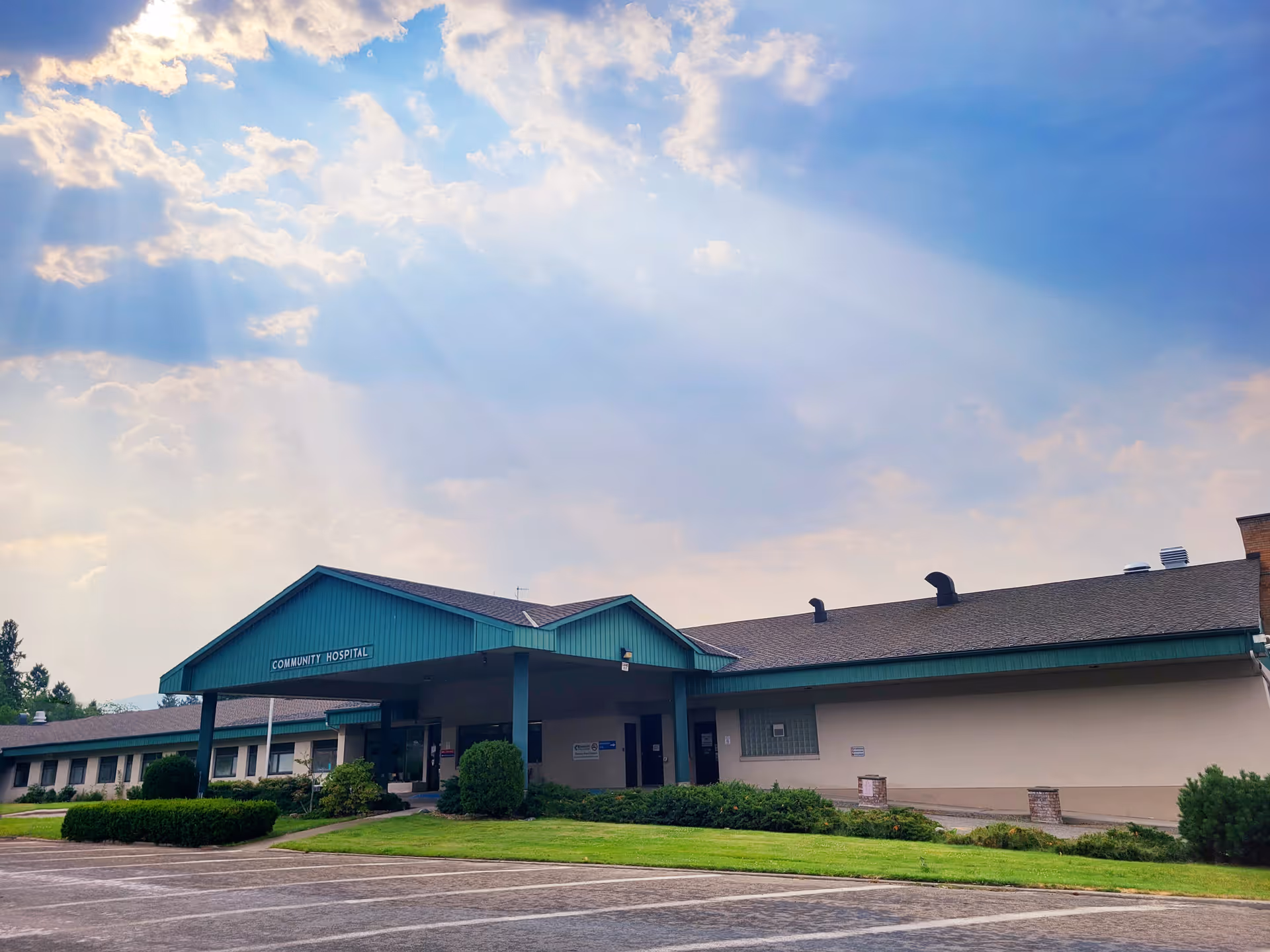 Front exterior of a one-story community hospital building with a green roof and entrance canopy, lawn and parking in the foreground beneath a partly cloudy sky.