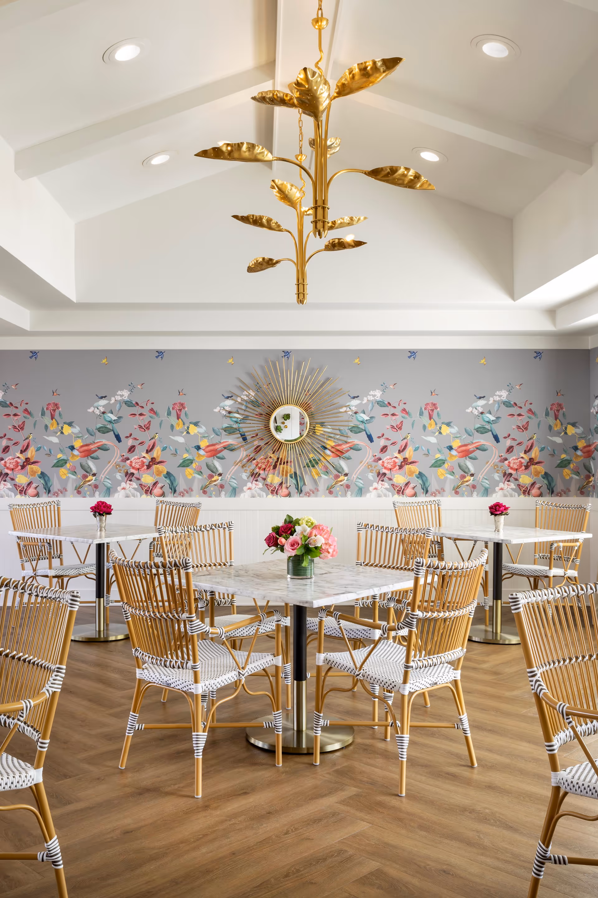 A bright dining room with several square marble-top tables surrounded by wicker chairs with white and natural wood tones. Each table has a small vase with colorful flowers. The walls feature a decorative floral and bird wallpaper with a sunburst mirror in the center. A gold chandelier with leaf-shaped elements hangs from the white vaulted ceiling. The floor is a warm wood with a herringbone pattern.