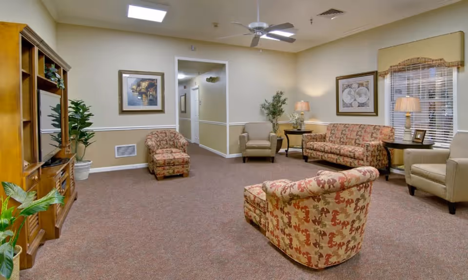 A cozy living room area in a senior living facility with patterned armchairs and a matching sofa, beige walls with white trim, framed artwork, potted plants, a wooden entertainment center with a TV, a ceiling fan, and table lamps providing warm lighting.