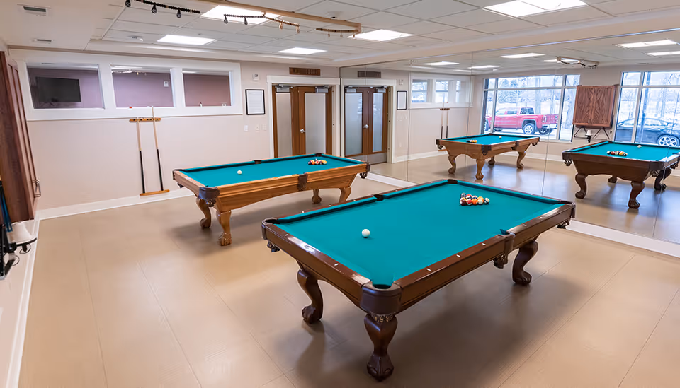 A recreational room with two wooden pool tables with green felt tops, one in the foreground and one in the background. The room has beige walls, a large mirror on one side, and windows showing parked cars outside. Pool cues are mounted on the wall, and there is a TV screen on the left wall.