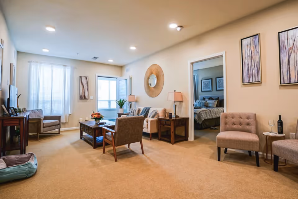 A cozy living room area with beige walls and carpet, featuring a sofa with pillows, two armchairs, a wooden coffee table with a flower arrangement, a side table with a lamp, and a TV on a stand. There is a doorway leading to a bedroom with a bed and framed artwork on the walls. The room is well-lit with recessed ceiling lights and natural light from a window with sheer curtains.