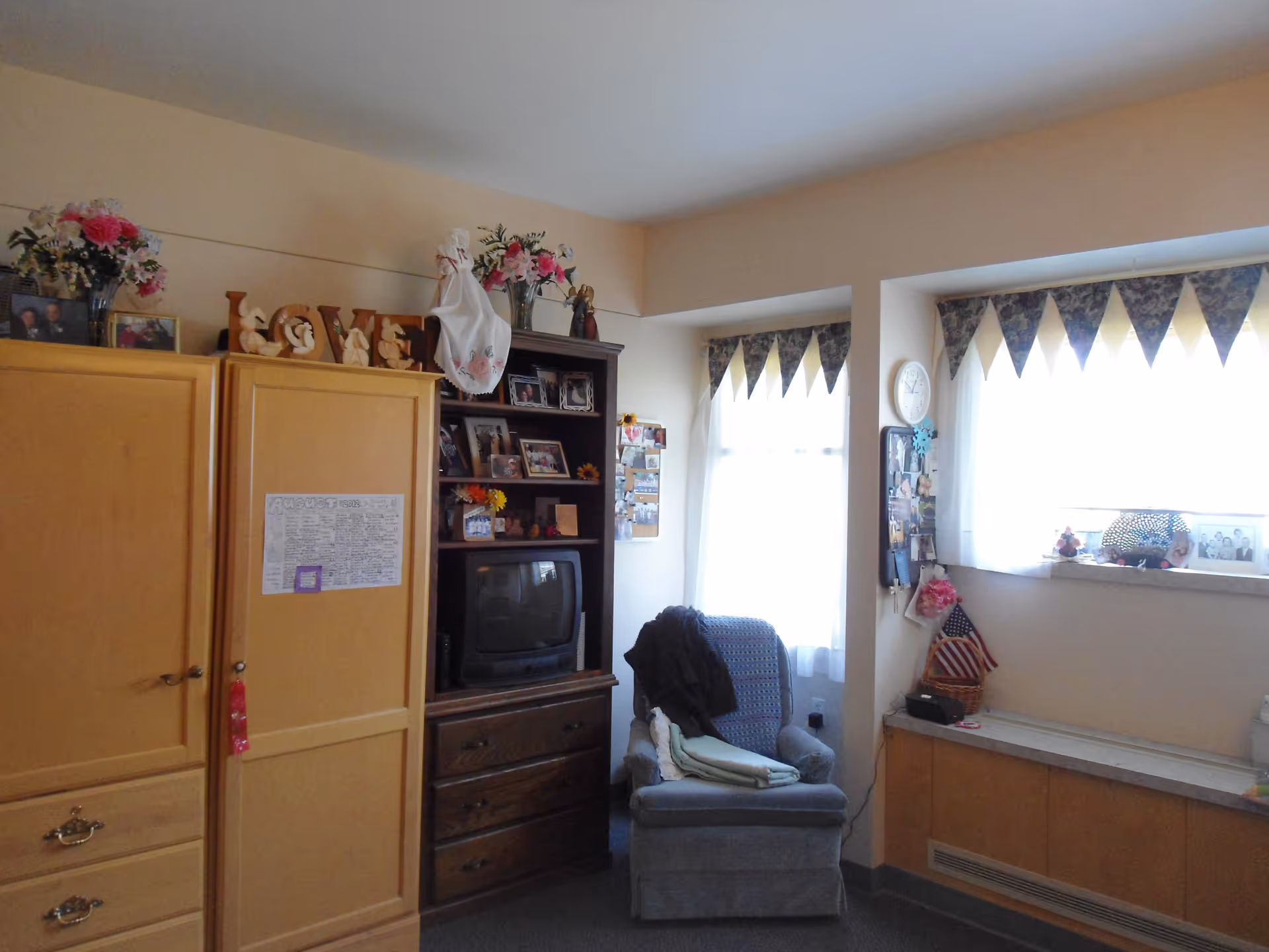 A cozy room with a wooden wardrobe, a dark wooden shelving unit holding a small TV and framed photos, a comfortable armchair with folded blankets, and two windows with patterned valances letting in natural light. The room is decorated with flowers, an American flag, and various personal items.