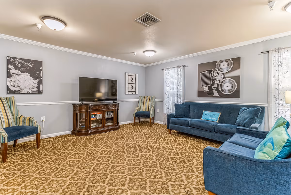Bright common living room with blue sofas, striped armchairs, a TV on a wooden console, and patterned carpet.
