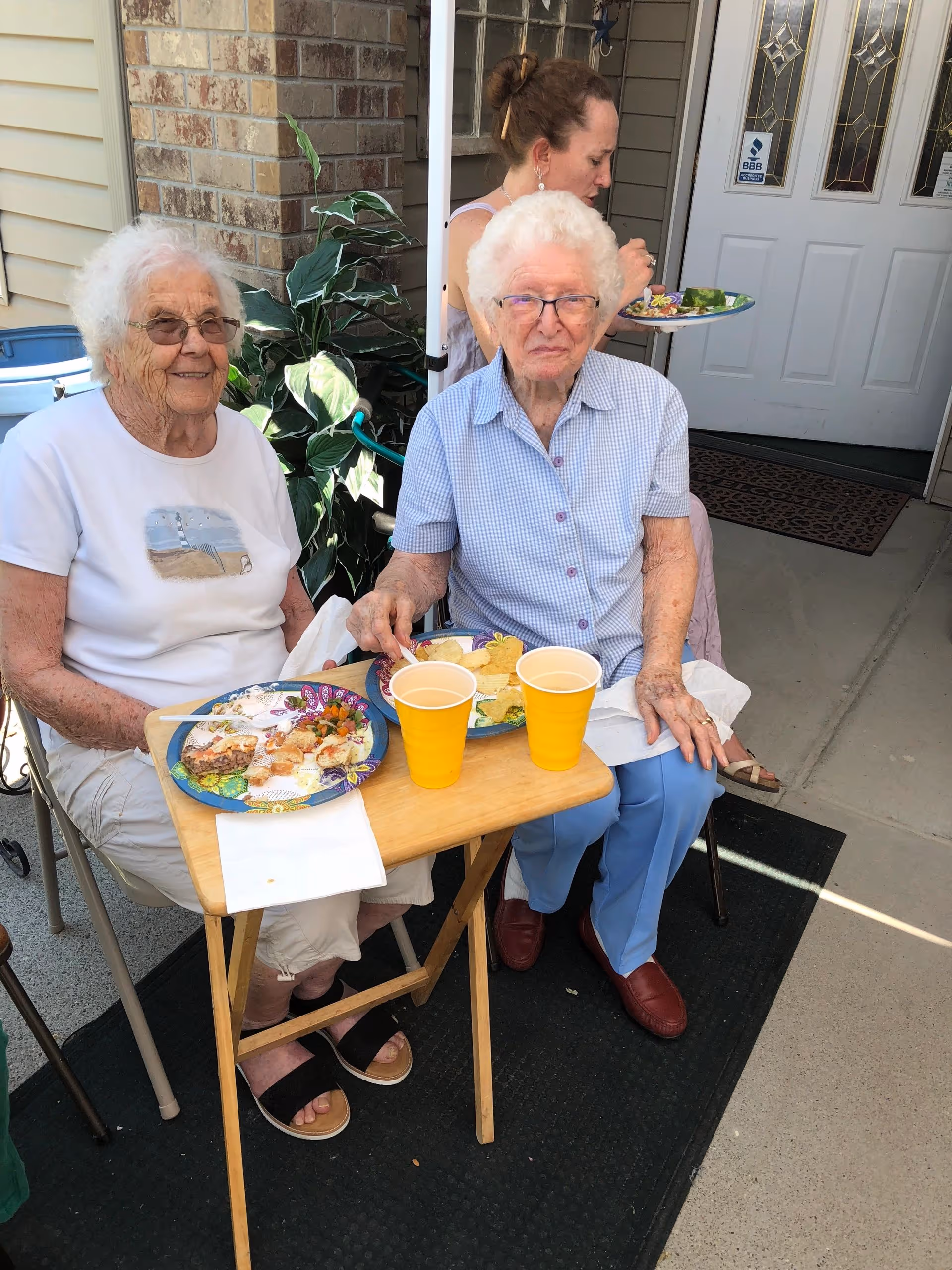 Two elderly women sitting outside at a small wooden table with plates of food and yellow cups in front of them. One woman is wearing a white t-shirt and glasses, the other is wearing a light blue checkered shirt and glasses. A younger woman in the background is holding a plate of food near a white door with decorative glass panels.