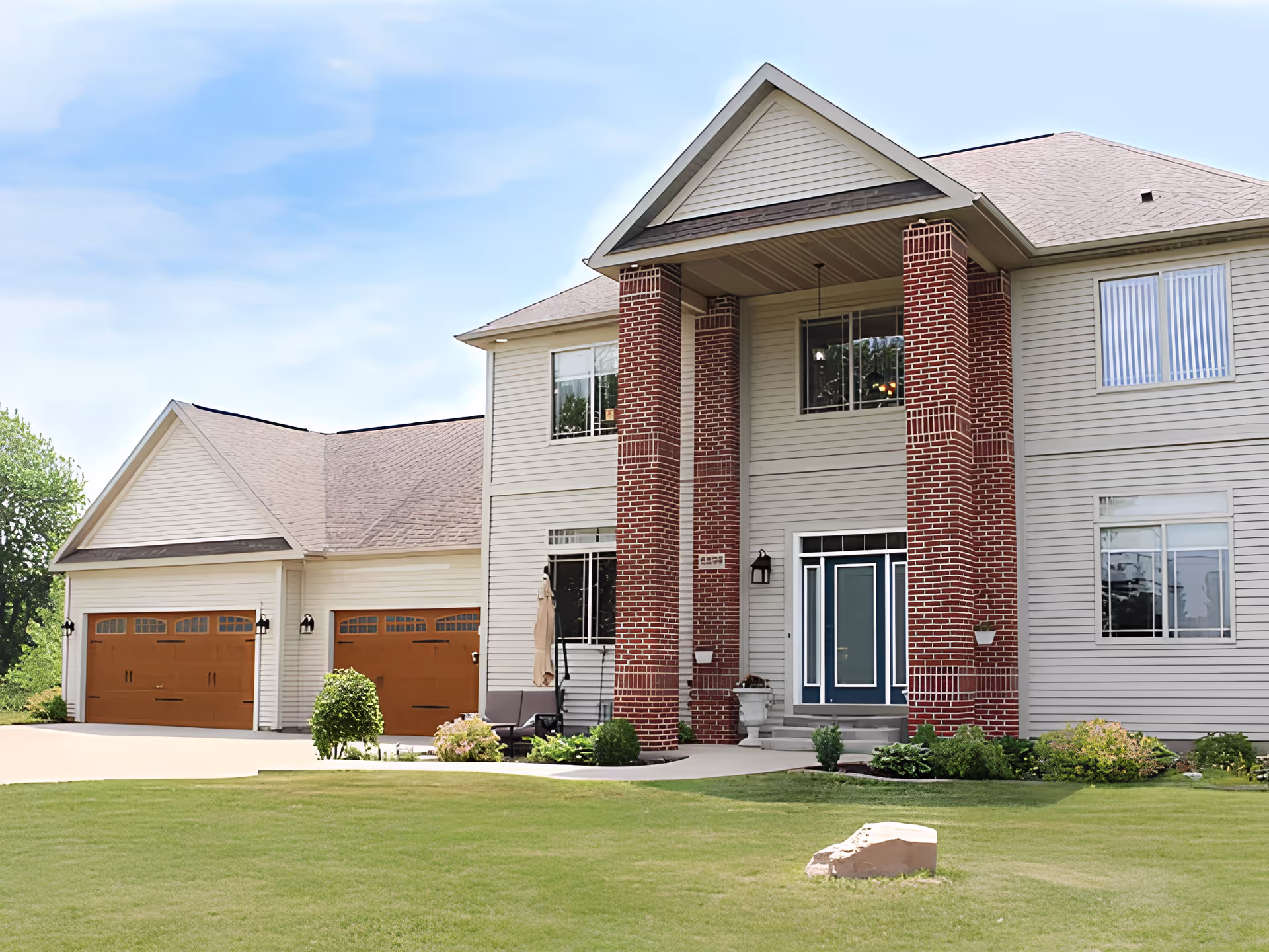 Front exterior view of a two-story residential building with beige siding and red brick columns. The house features a blue front door, multiple windows, a double garage with wooden doors, and a well-maintained green lawn with some shrubs and a small rock in the foreground under a partly cloudy sky.