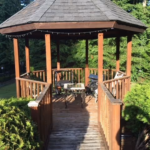 Wooden gazebo with a shingled roof situated in a lush green garden. Inside the gazebo, there is a glass-top table and several chairs. The gazebo is accessed by a wooden ramp with railings on both sides. Surrounding the gazebo are dense green trees and bushes.