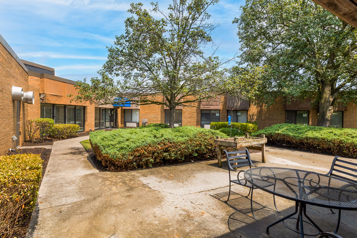 Outdoor courtyard area at Autumn Lake Healthcare at Patuxent River featuring a concrete pathway, green bushes, trees, and a black metal table with chairs under a blue sky.