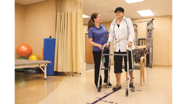 A rehabilitation room with exercise equipment and therapy balls. A healthcare worker in a blue shirt assists an elderly woman using a walker as she practices walking along a marked path on the floor.