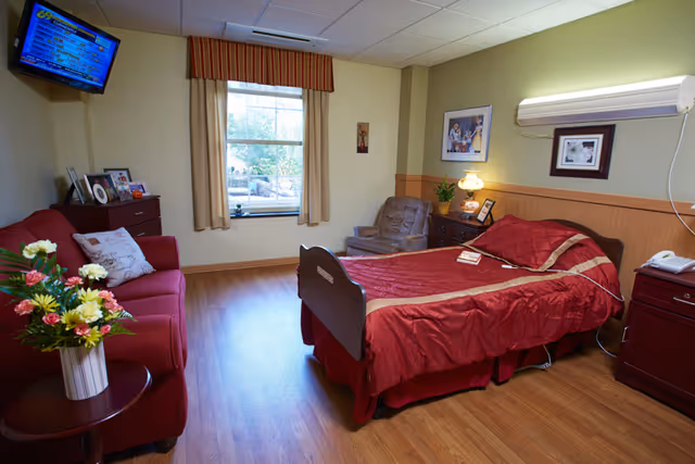 A cozy senior living bedroom with a single bed covered in a red and gold bedspread, a red sofa with pillows, a small round table with a vase of flowers, a window with beige curtains, a wall-mounted TV, a comfortable armchair, and wooden flooring. The room has soft yellow walls with wood paneling and framed pictures on the walls.