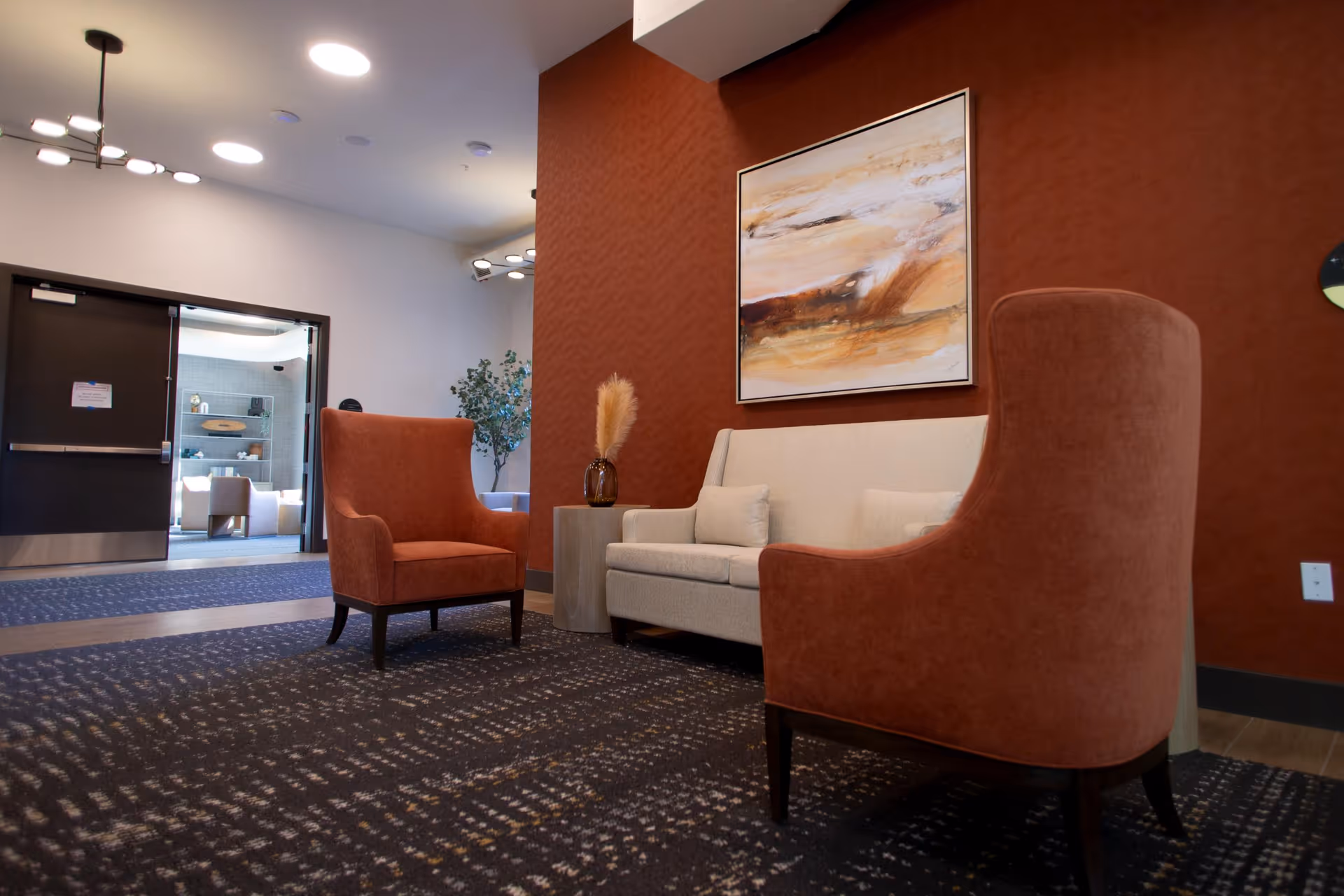 A cozy seating area in a retirement community featuring two rust-colored armchairs and a light beige sofa against a textured rust-colored wall. A modern abstract painting hangs above the sofa, and a small side table with a decorative vase and dried pampas grass is placed between the sofa and one armchair. The floor is carpeted with a patterned design, and a doorway leads to another room with additional seating and shelves.