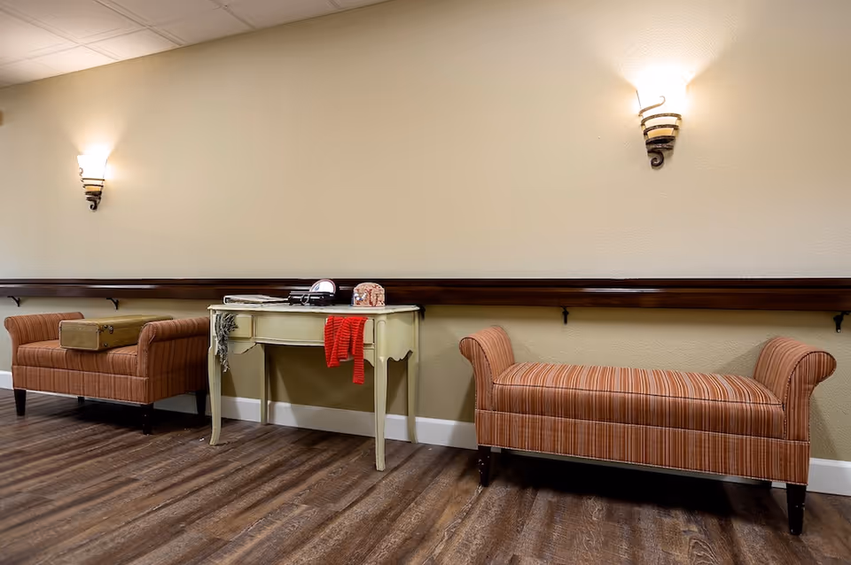 A hallway area with two striped upholstered benches on either side of a small cream-colored table. The table has a red scarf draped over one side and some small items on top. The walls are beige with two wall-mounted light fixtures above the benches, and the floor is a dark wood laminate.