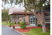 Exterior view of a brick building with a striped red and white awning over the entrance. There are potted plants and landscaping with mulch and green grass around the entrance area. Trees are visible near the building.