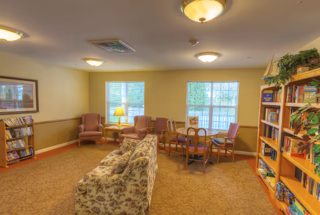 A cozy senior living common room with a floral sofa, armchairs, a round table with chairs, bookshelves and windows letting in daylight.