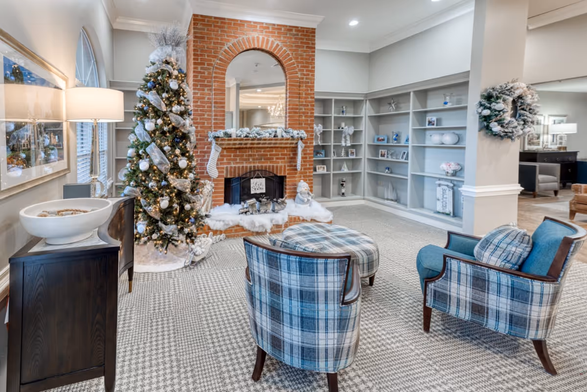 A cozy living room area decorated for Christmas with a tall Christmas tree adorned with silver ornaments and ribbons next to a brick fireplace. The fireplace mantel is decorated with garland and stockings. In front of the fireplace are two plaid upholstered chairs and a matching ottoman. Built-in shelves with decorative items line the wall, and a wreath hangs on a pillar. The room has soft lighting and a patterned carpet.