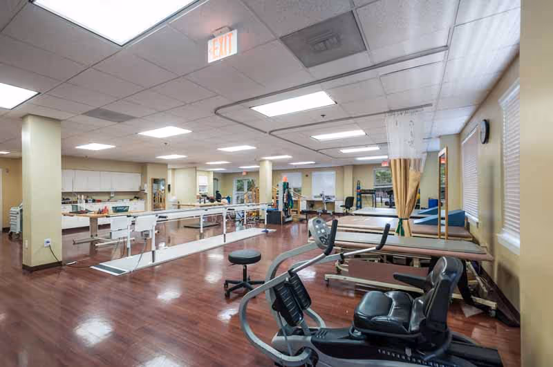 Spacious rehabilitation gym with exercise equipment, parallel bars, and treatment tables in a senior living facility.