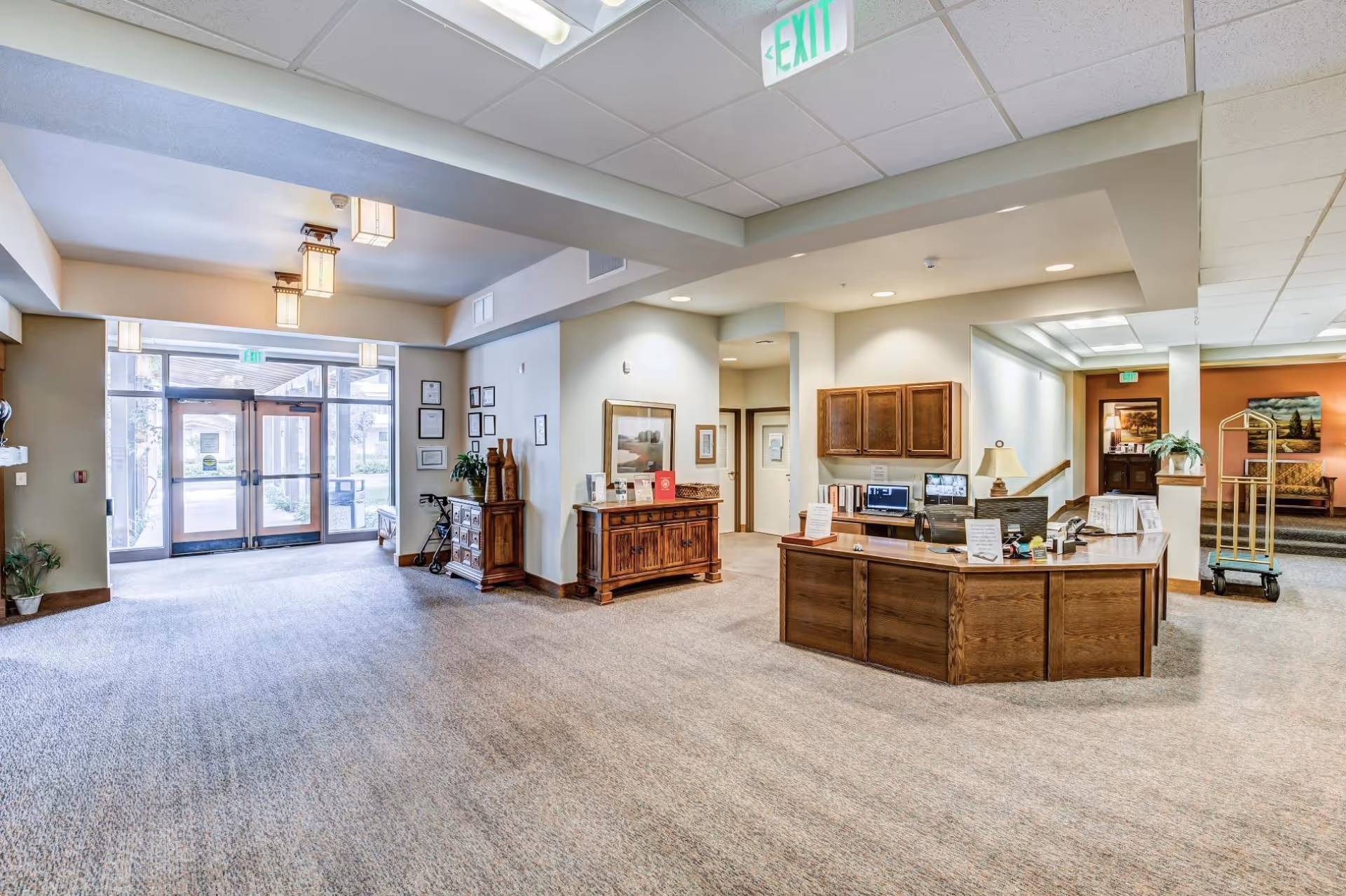 Spacious and well-lit assisted living facility lobby with a wooden reception desk, carpeted floor, decorative wooden furniture, framed artwork on the walls, and glass double doors leading outside. There is an exit sign above the doors and a luggage cart near a seating area in the background.