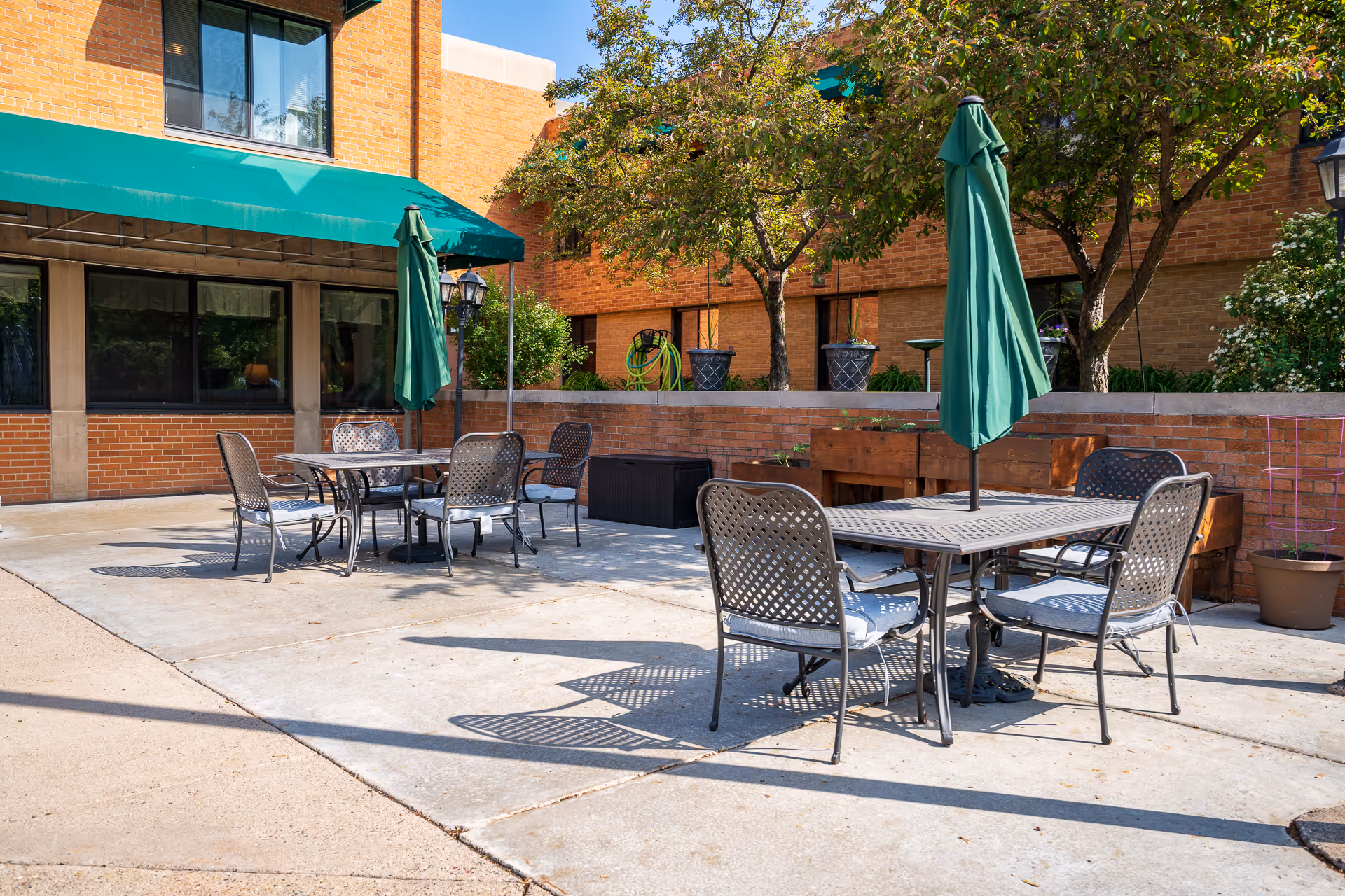Outdoor patio area with metal tables and chairs, each table having a closed green umbrella. The patio is surrounded by brick buildings and trees with green foliage. There are potted plants and a garden hose visible in the background.