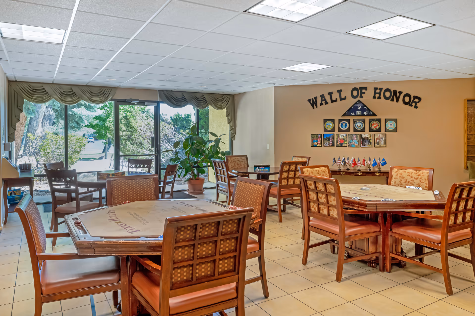 A well-lit common area with multiple wooden tables and chairs arranged for group activities. The room features a 'Wall of Honor' display with military insignias, photos, and small flags. Large windows and a glass door provide a view of greenery outside.