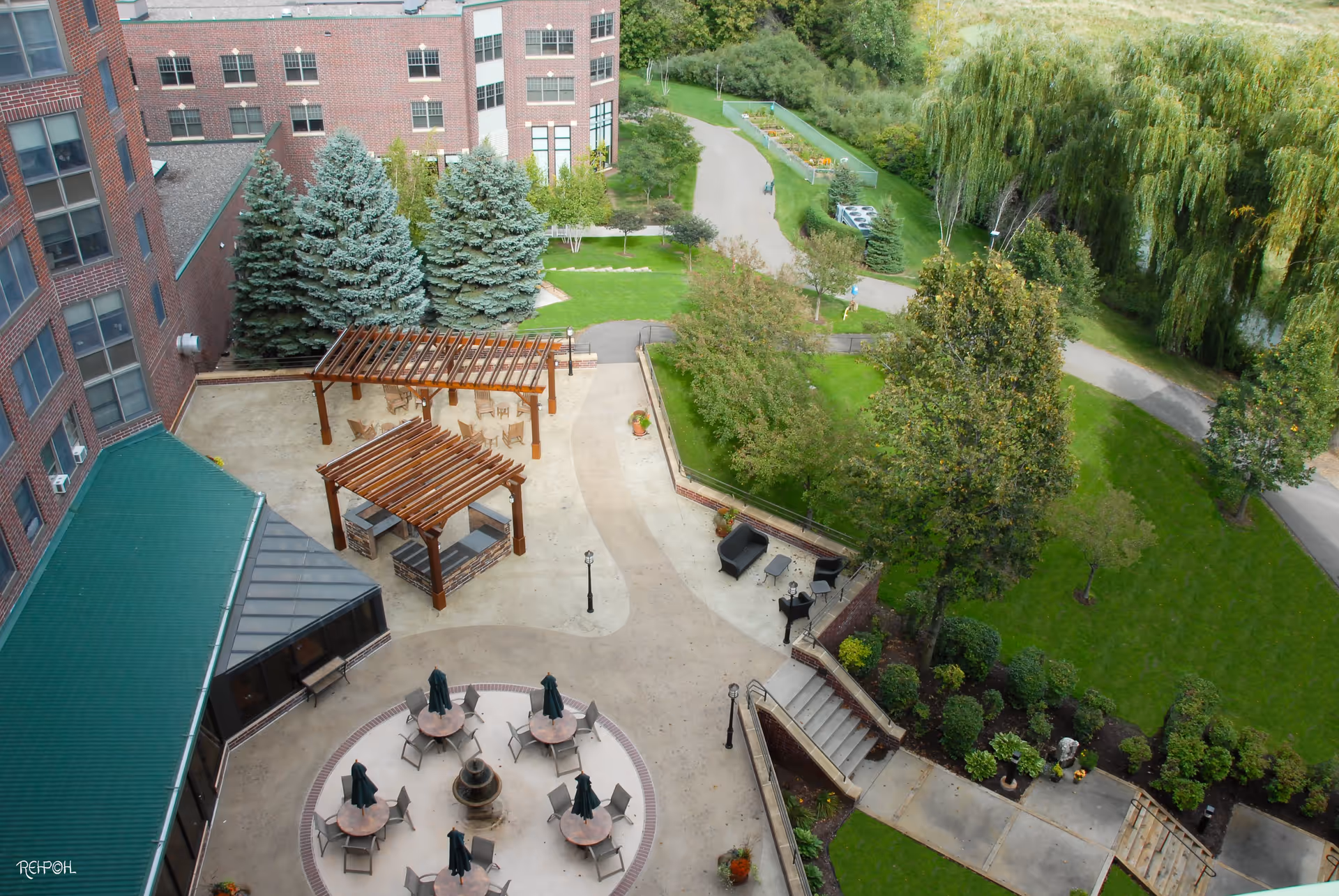 Aerial view of an outdoor patio area at The Glenn Hopkins facility featuring multiple round tables with chairs and closed umbrellas, two wooden pergolas with seating underneath, a small fountain in the center of the patio, and surrounding greenery including trees, shrubs, and a walking path.