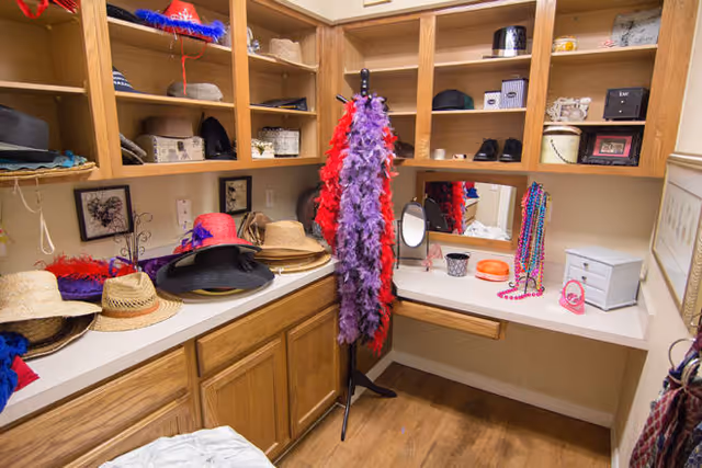 A dressing room with wooden shelves and cabinets filled with various hats, feather boas, costume jewelry, and small decorative items. There is a countertop with a mirror, a small vanity mirror, and a few personal accessories. The floor is wooden, and the room has a cozy, organized feel.