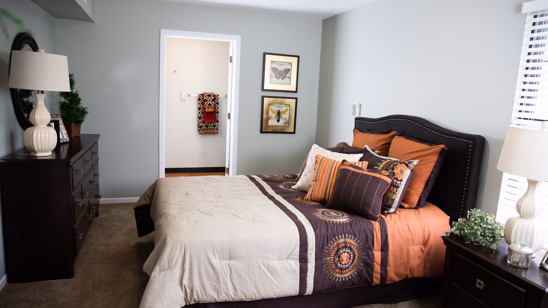 A neatly made bed with patterned orange, brown, and beige bedding in a bedroom. The room features a dark wooden dresser with a lamp and a plant on top, a nightstand with a lamp and a small plant, and two framed pictures on the wall. There is an open doorway leading to a bathroom with towels hanging on the wall.
