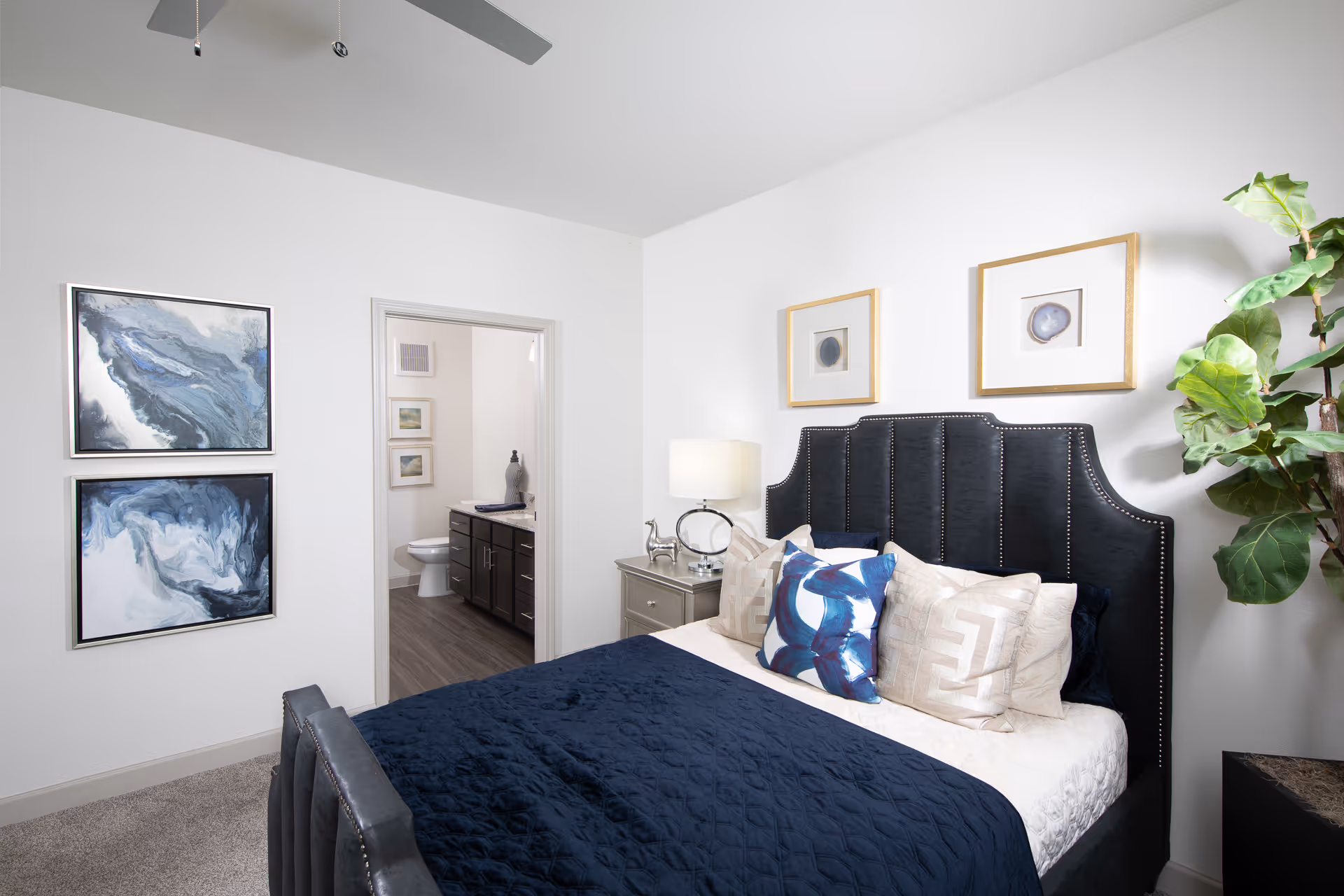 A modern bedroom with a dark upholstered headboard, a bed with blue and beige pillows and a blue quilt, a nightstand with a lamp and decorative items, two framed artworks above the bed, two abstract paintings on the wall, a potted plant in the corner, and a view into an adjoining bathroom with a toilet and vanity.