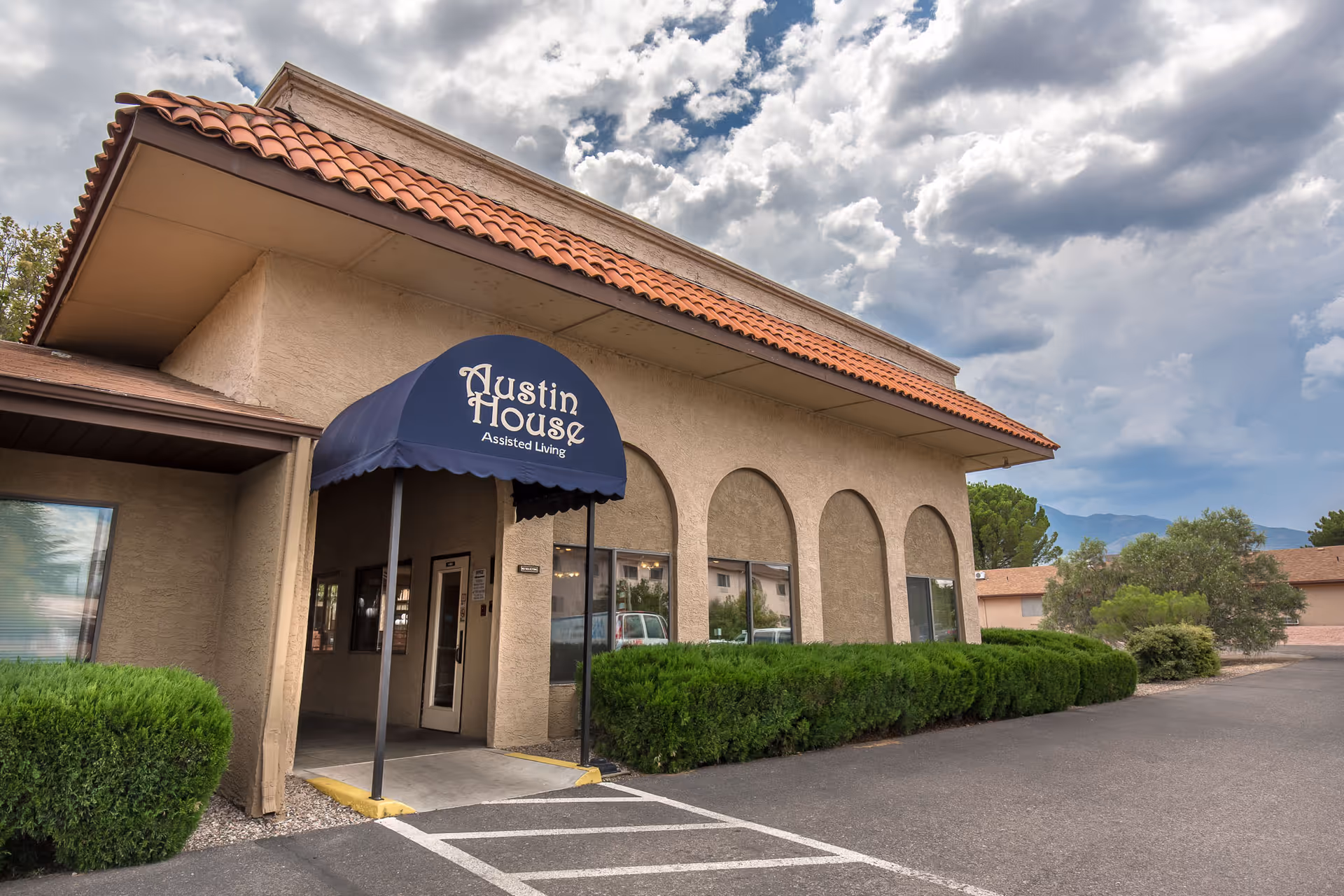 Exterior view of a beige stucco building with a red tile roof and a blue awning that reads 'Austin House Assisted Living'. There are bushes along the front of the building and a parking area in front. The sky is partly cloudy with mountains visible in the background.