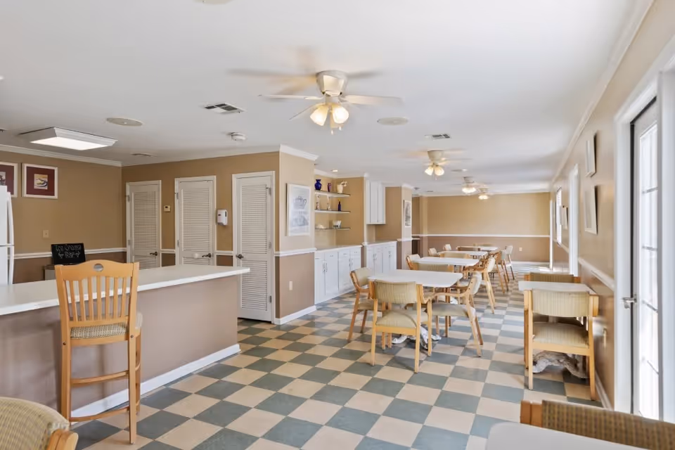 Communal dining room with checkerboard tile floor, multiple tables and chairs, a serving counter, and ceiling fans.