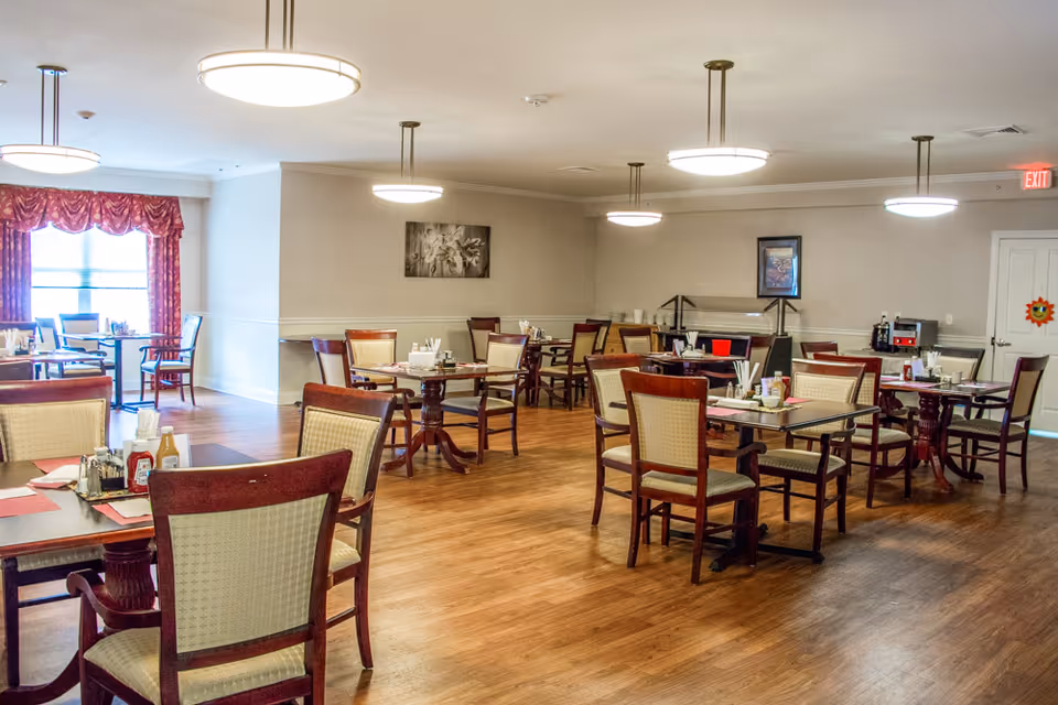 A spacious dining room with multiple wooden tables and chairs arranged neatly. The room has wooden flooring, beige walls, and several round ceiling lights. There are condiment holders on the tables and a window with red curtains letting in natural light.