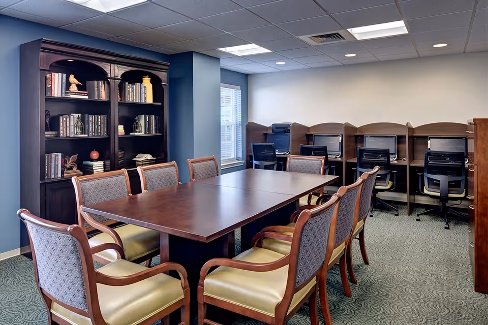 A room with a large wooden table surrounded by eight upholstered chairs. Behind the table is a dark wooden bookshelf filled with books and decorative items. Along the back wall, there are four computer workstations with chairs, each separated by wooden dividers. The room has blue walls, a window letting in natural light, and a carpeted floor.