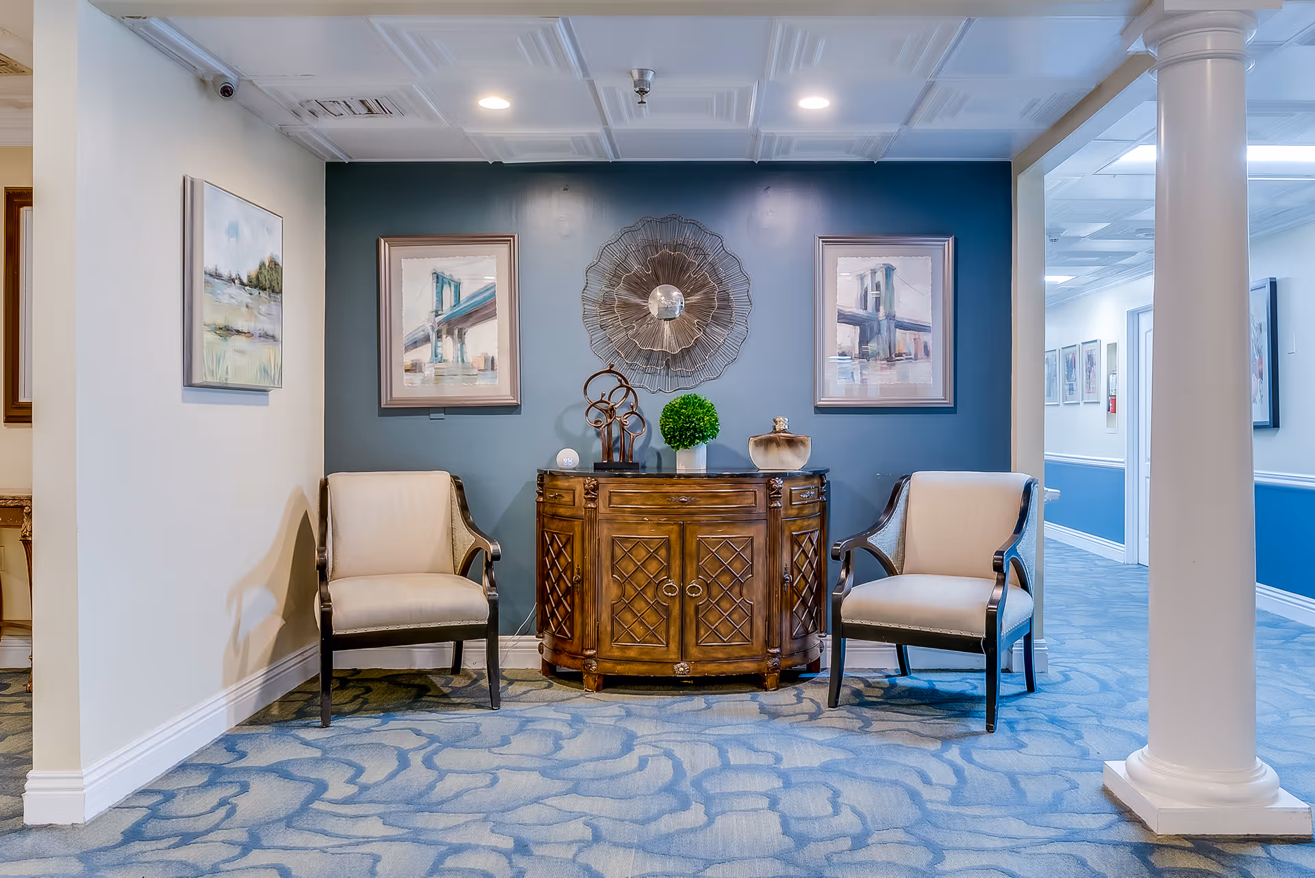 A cozy seating area in a senior living facility featuring two beige armchairs on either side of a wooden cabinet with decorative items on top, including a small green plant and a sculpture. The wall behind is painted blue and adorned with two framed paintings of a bridge and a decorative metal wall art piece in the center. The floor is carpeted with a blue patterned design, and a white column is visible on the right side.
