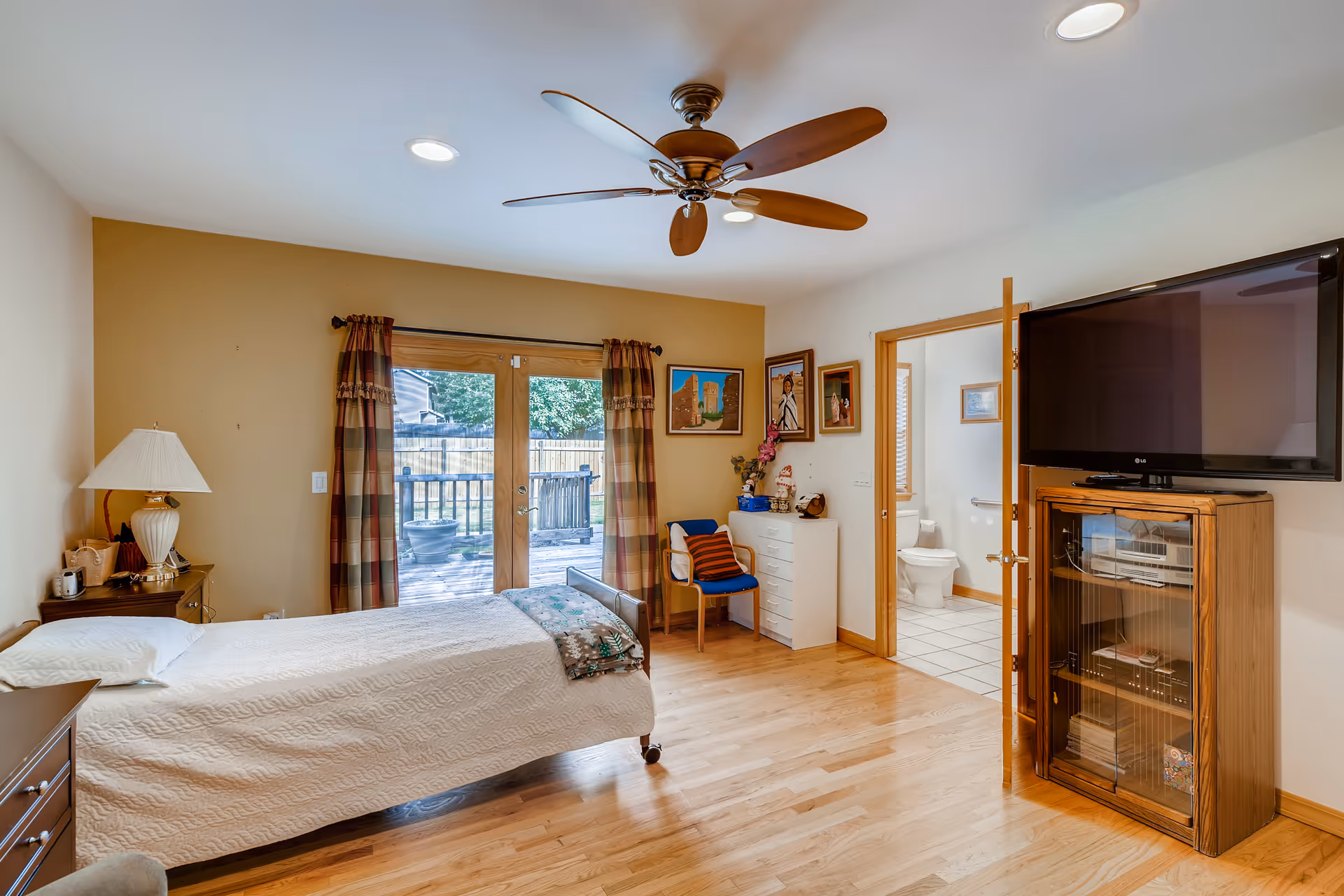 A senior living bedroom with a single bed covered in a white quilt, a wooden nightstand with a lamp, a ceiling fan, a large flat-screen TV mounted on a wooden cabinet, and a door leading to a bathroom. The room has wooden floors, beige walls, and glass doors with plaid curtains opening to an outdoor deck.