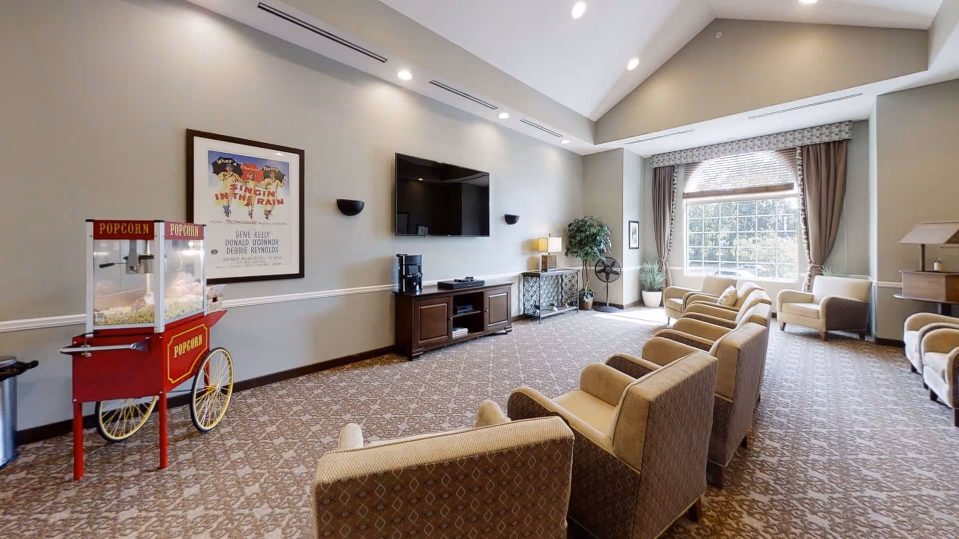 Spacious communal lounge with rows of upholstered chairs facing a wall-mounted TV, a popcorn cart, and a large window.
