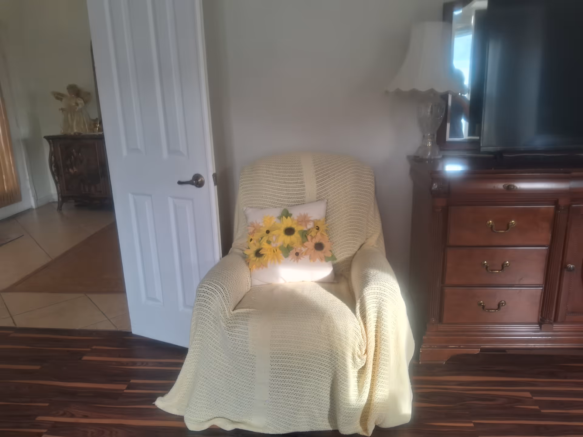 A cozy armchair covered with a pale yellow knitted throw and a sunflower-decorated pillow sits between a partially open white door and a wooden TV dresser on dark hardwood floors.