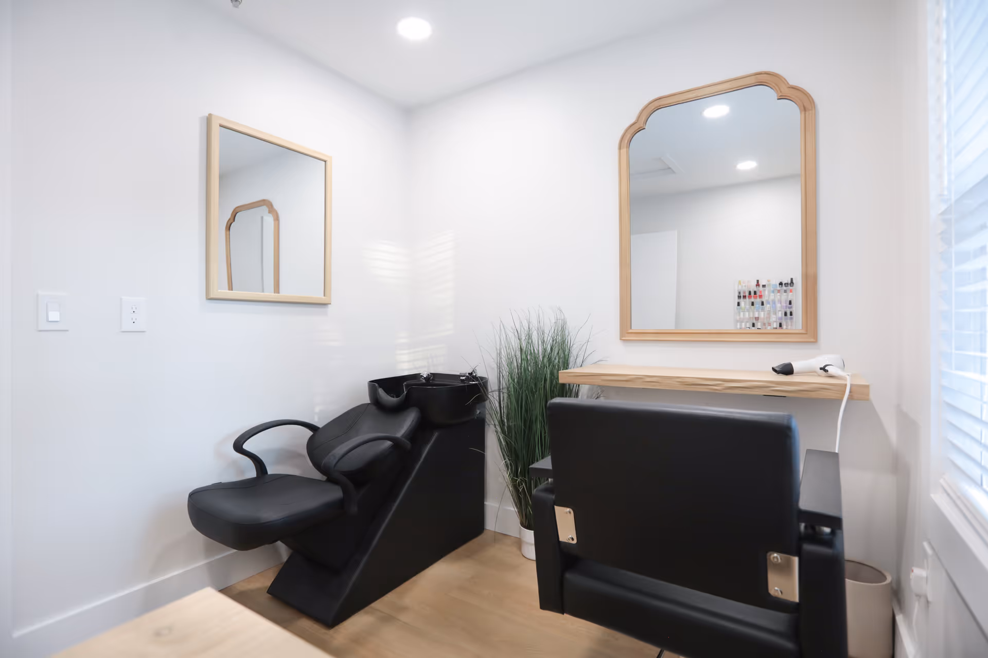 A small hair salon area with a black salon chair and a black shampoo sink. There are two wooden framed mirrors on the white walls, a wooden shelf with a hair dryer on it, and a potted plant in the corner. The room has bright lighting and a window with blinds.