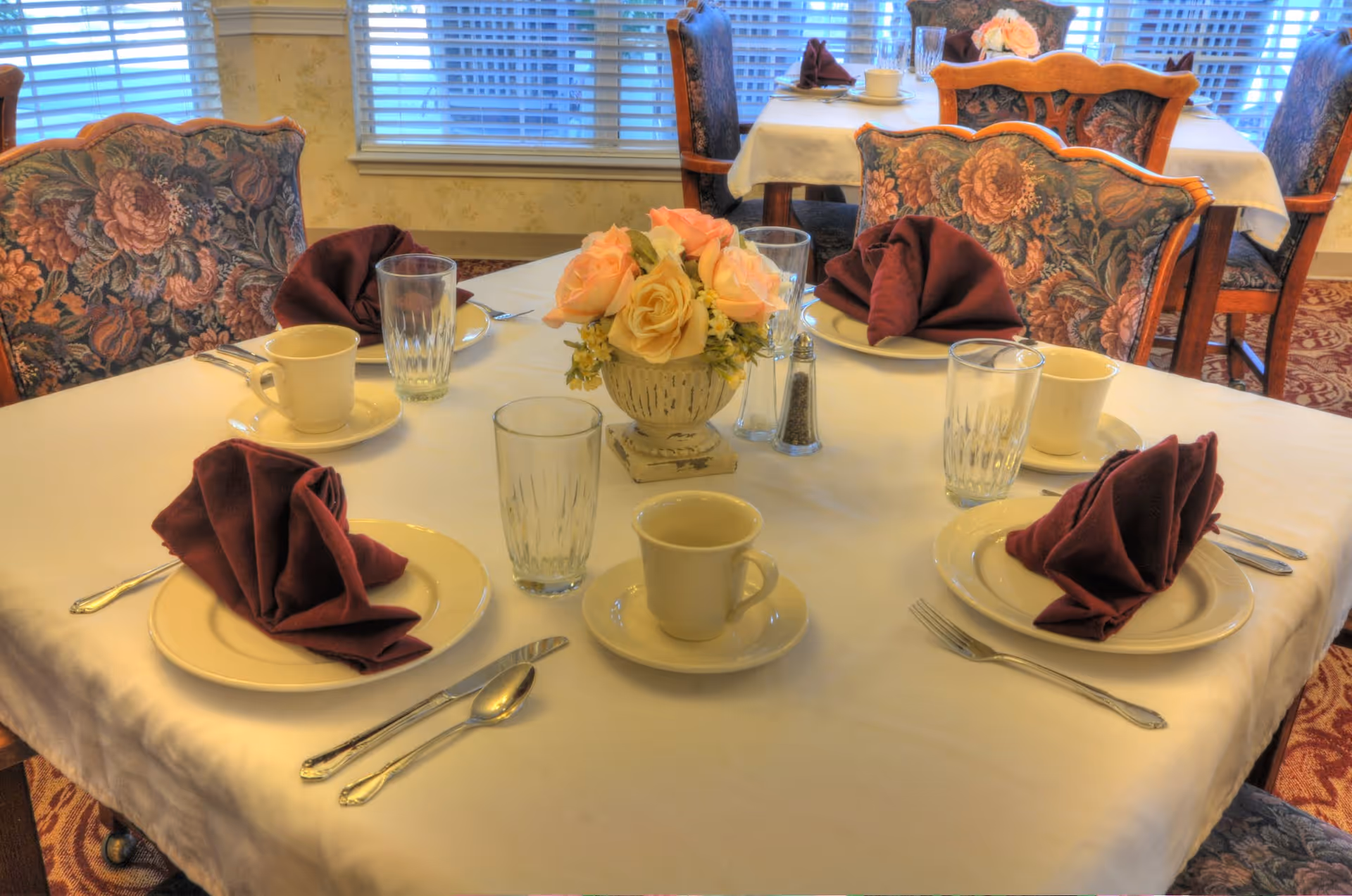 A dining table set for four with white tablecloth, beige plates, maroon folded napkins, clear drinking glasses, cream-colored coffee cups with saucers, silverware, and a floral centerpiece with pink and yellow roses. The background shows floral upholstered chairs and windows with blinds.