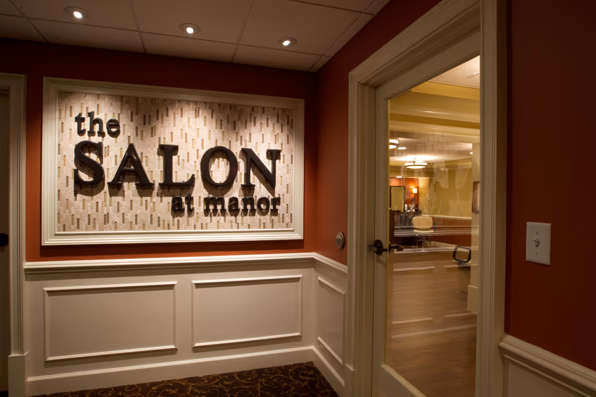 Hallway entrance to "The Salon at Manor" with a framed sign on a red wall and a glass door showing the salon interior.