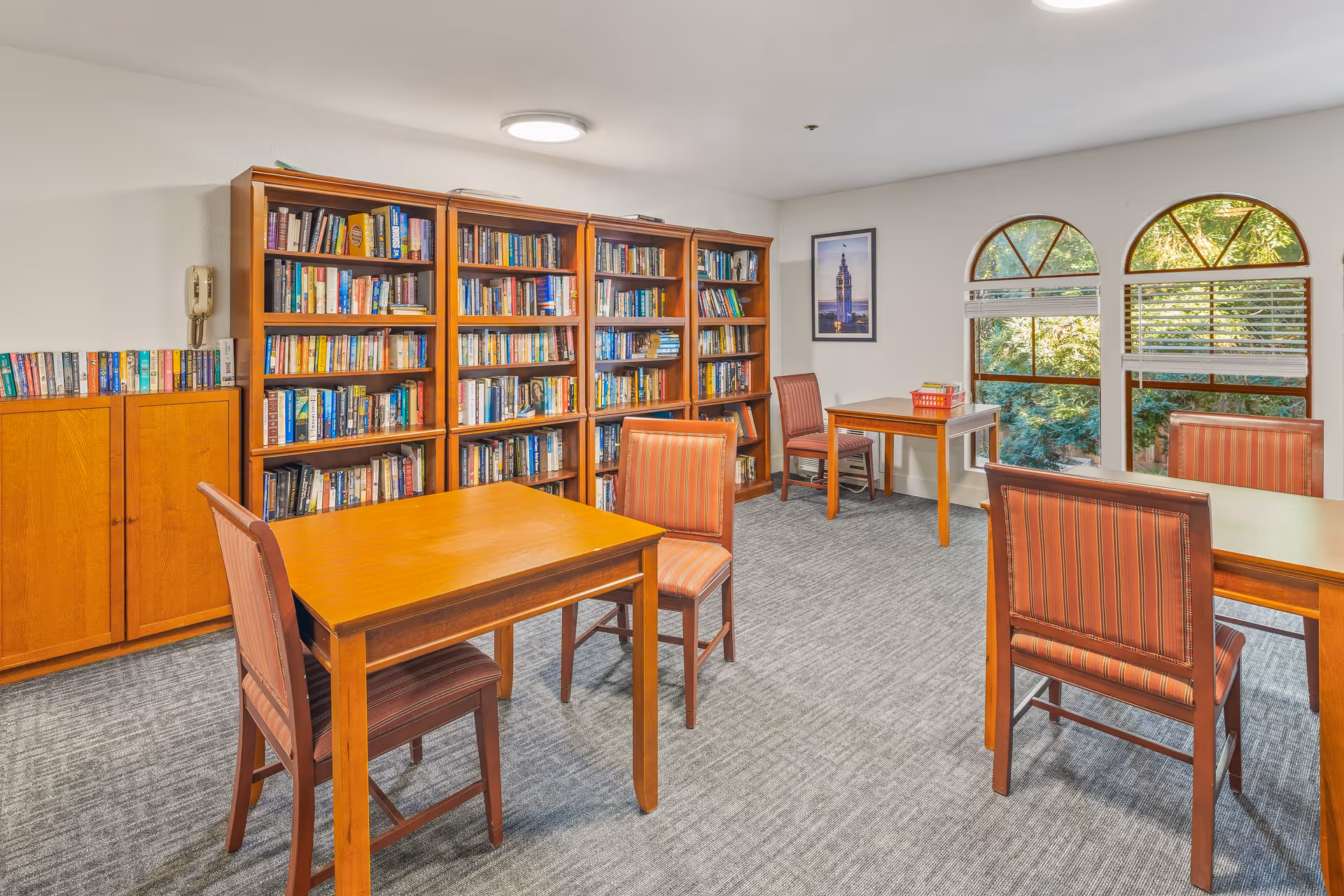 A well-lit room with wooden tables and chairs featuring red striped upholstery. There are three large wooden bookshelves filled with books against one wall, and a wooden cabinet with more books on top. The room has carpeted flooring, two arched windows with blinds partially open showing greenery outside, and a framed picture on the wall.