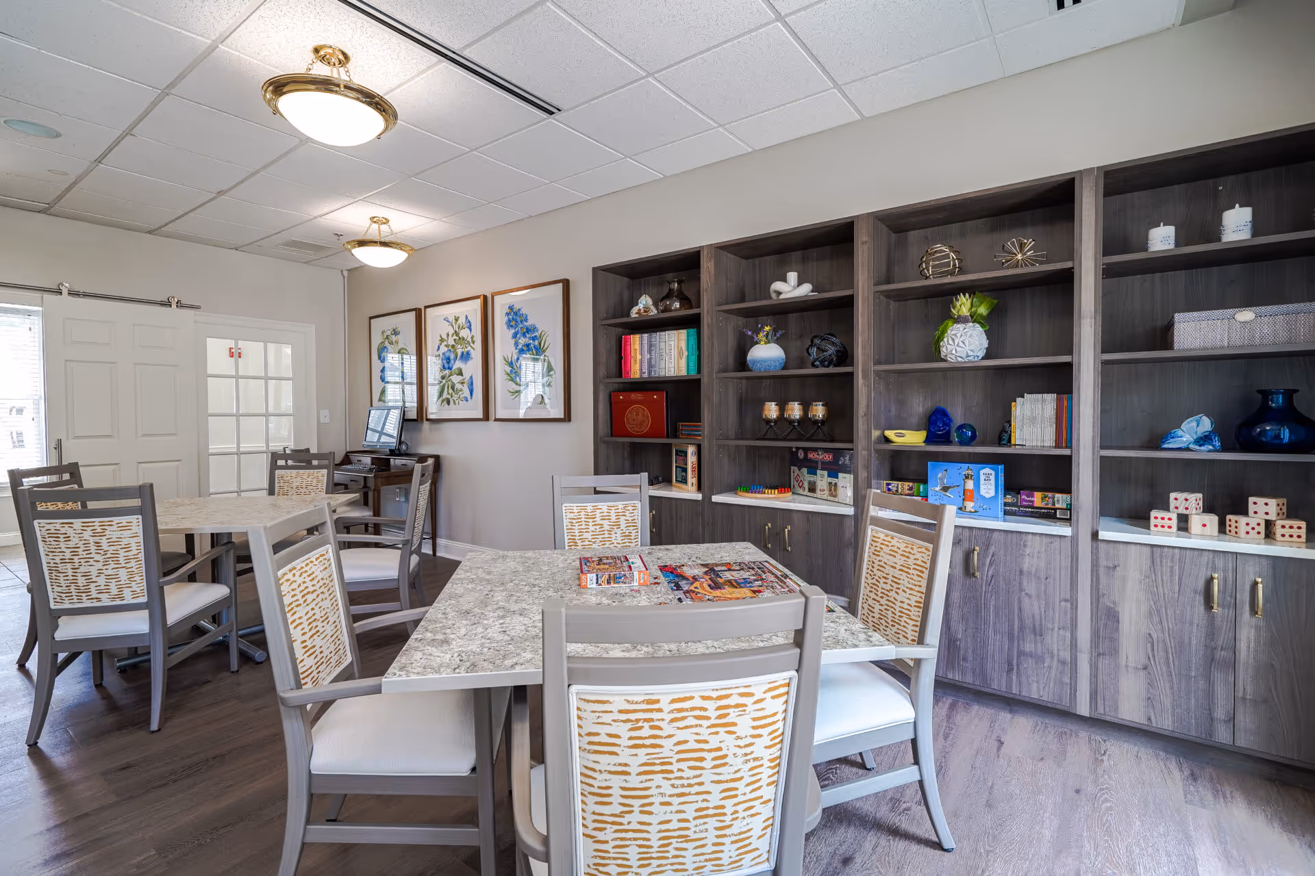 Bright communal activity room with several tables and chairs and a wall of shelves filled with books, games, and decor.