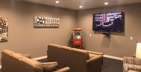 A cozy living room with two brown sofas facing a wall-mounted flat-screen TV showing a courtroom scene. There is a popcorn machine in the corner and a decorative theater-themed sign on the wall. A patterned armchair and a lamp are partially visible on the right side.