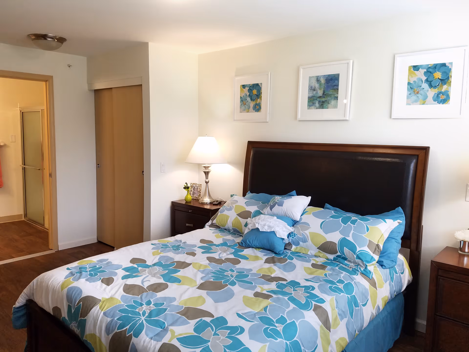 A bedroom with a bed featuring a floral patterned bedspread in shades of blue, green, and white. The bed has a dark wooden frame with a padded headboard. On either side of the bed are wooden nightstands, one with a lamp and a small vase with flowers, and the other with a decorative item. Three framed floral artworks hang on the wall above the bed. A doorway leads to a bathroom with a shower visible.