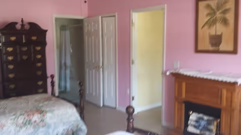 A bedroom with pink walls featuring a wooden bed with floral bedding, a tall dark wooden dresser, an open door leading to a bathroom, and a wooden fireplace with a framed palm tree picture above it.